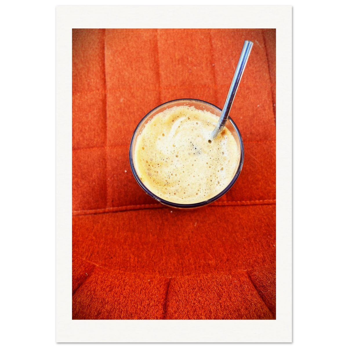 Kunstfotografi av latte i glass med metallsugerør, sett ovenfra mot rød designstol i tekstil. Kaffeskummet skaper abstrakte former som refererer til tradisjonen med å spå i kaffe.